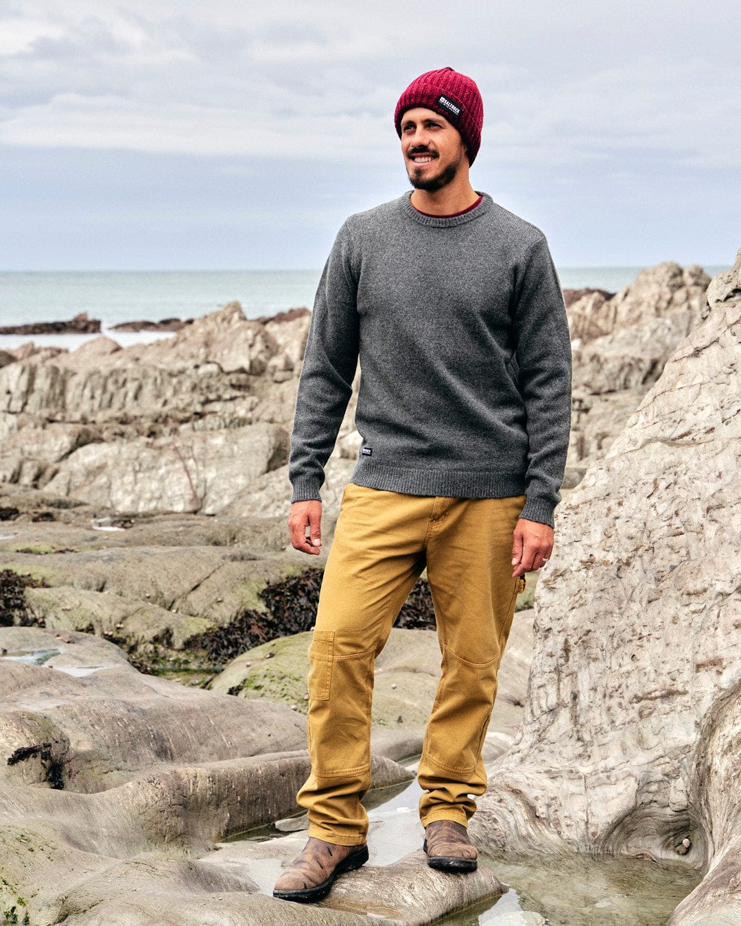 A man standing on rocks near the ocean, layering up with a Saltrock Bowen - Mens Knitted Crew - Dark Grey beanie and khaki pants.