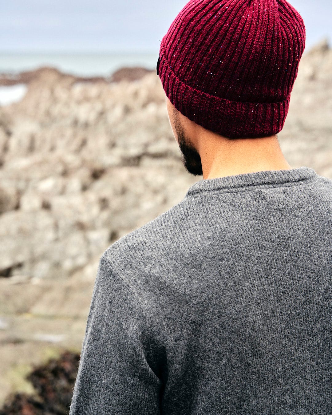 A man wearing a burgundy beanie, layering up against the cold breeze, gazes out at the ocean while wearing the Saltrock Bowen - Mens Knitted Crew in Dark Grey.