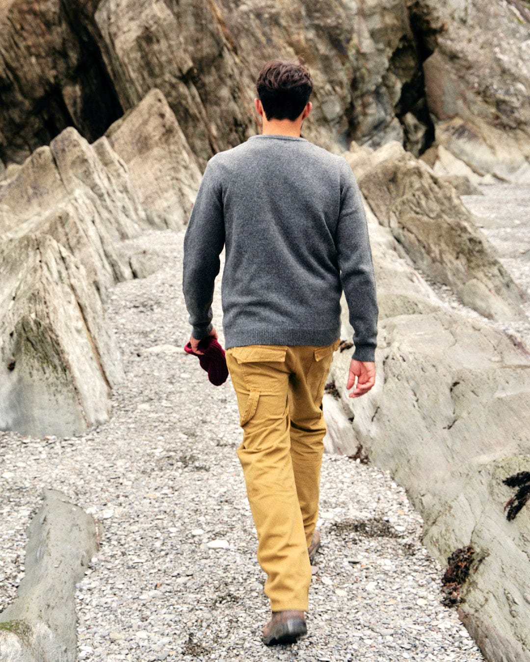 A man walking down a rocky path, carrying a bottle of water, showcasing the Saltrock branding on his clothing. He is dressed in the Saltrock Bowen - Mens Knitted Crew - Grey, perfect for layering during outdoor.