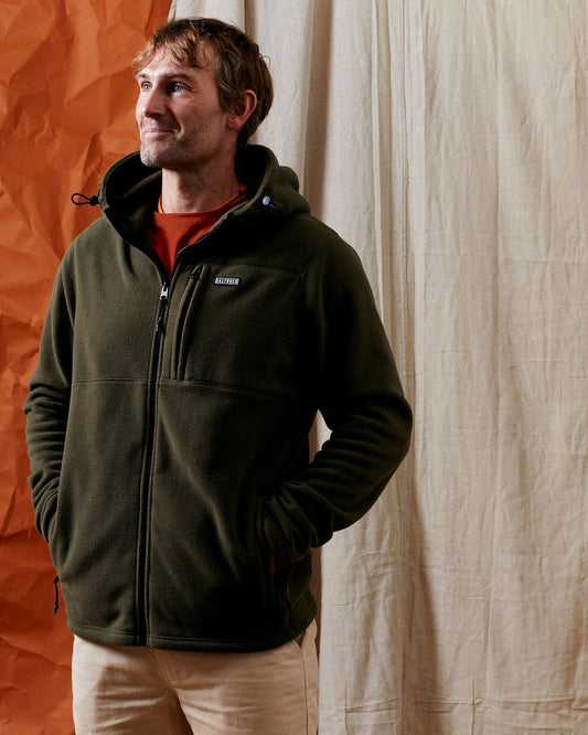 A person wearing the Saltrock Bradford Men's Recycled Hooded Fleece in green stands before a two-tone fabric backdrop, looking to the side.