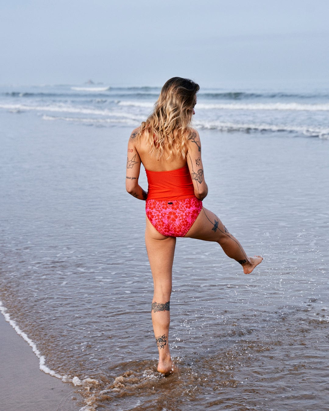 A woman in Saltrock's Caeley - Recycled Womens Hibiscus Bikini Bottoms in Pink walking into the ocean, her back to the camera, showing off various tattoos, with her foot kicking up water.