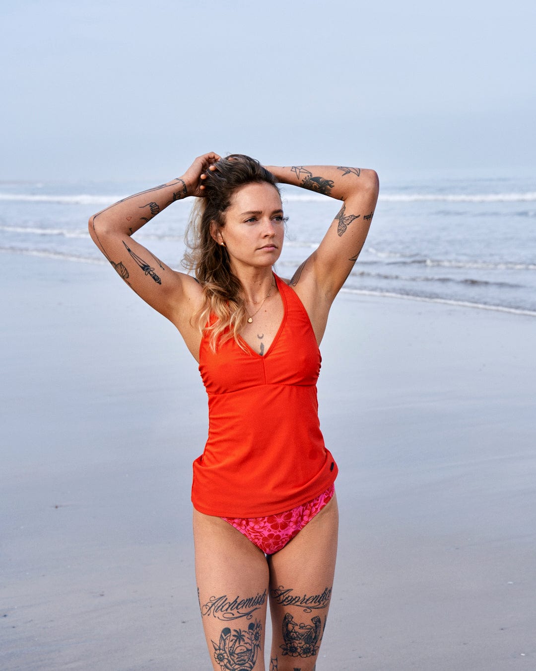 A woman in Caeley - Recycled Womens Hibiscus Bikini Bottoms - Pink by Saltrock with tattoos stands on a beach, hands lifting her hair, looking to the side.