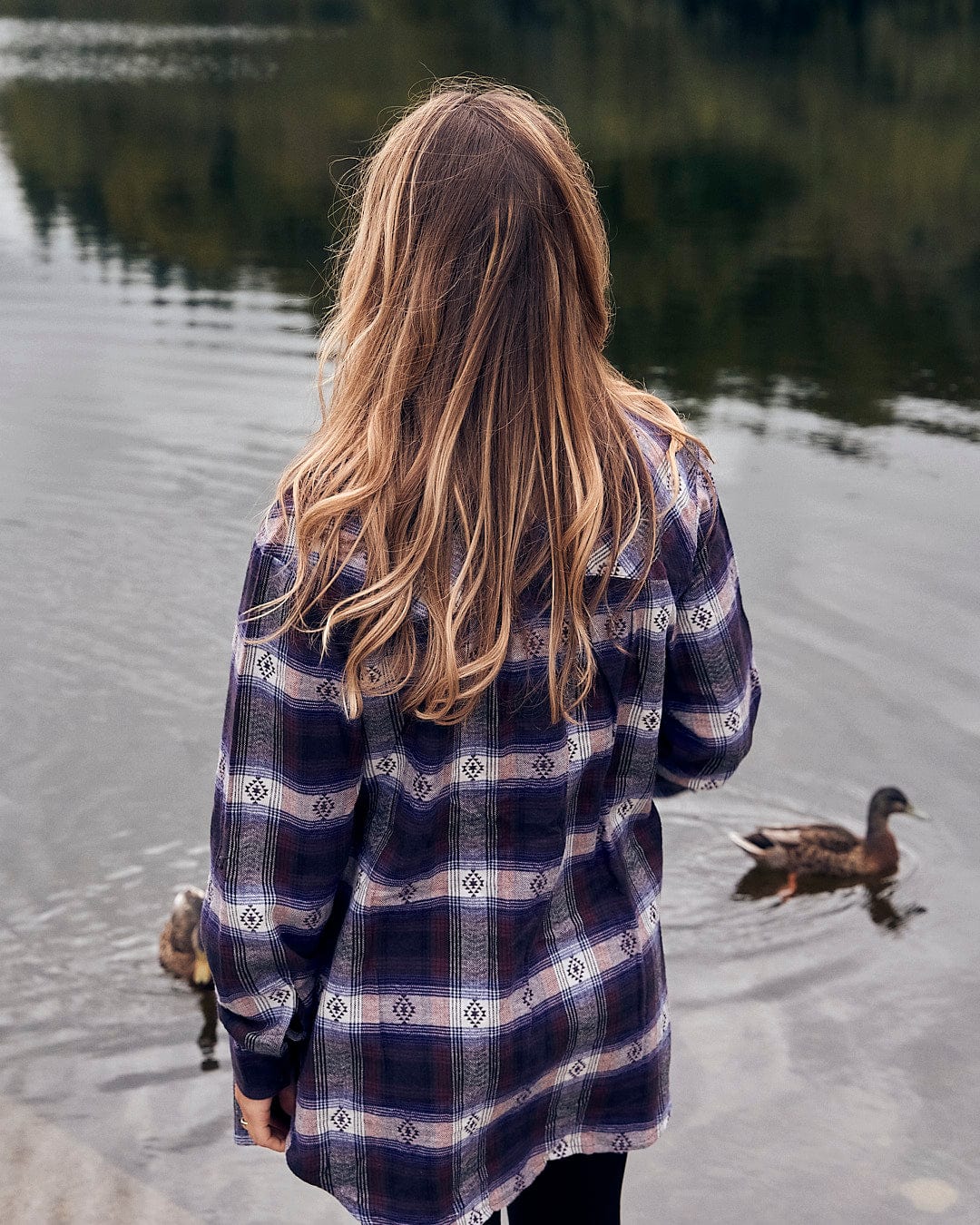 A woman wearing a Saltrock Camille - Womens Jaquard Check Boyfriend Shirt - Purple standing by a lake with ducks in the background.