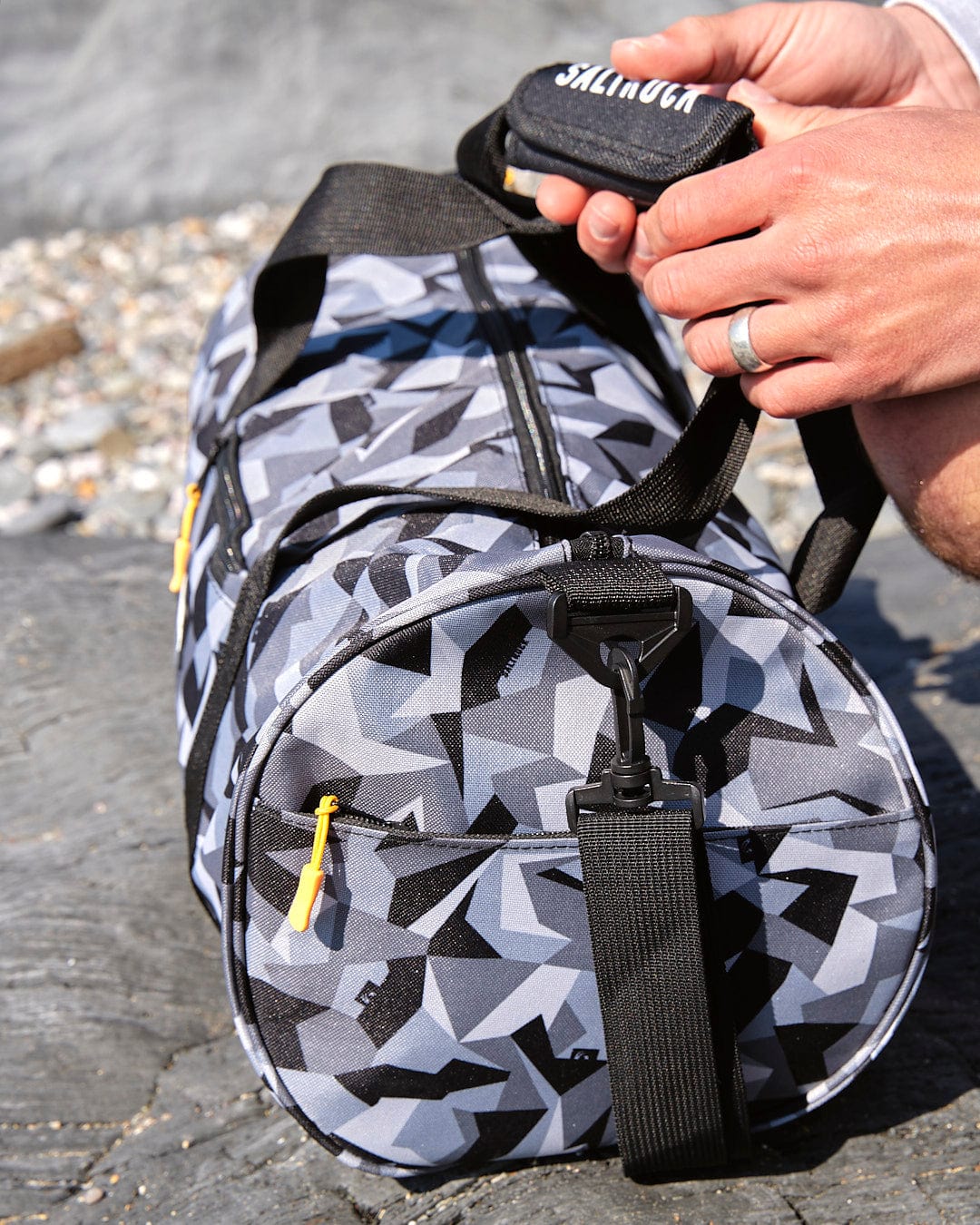 A person holding a Saltrock Camo Balboa - Hold-All Bag - Dark Grey on the beach.