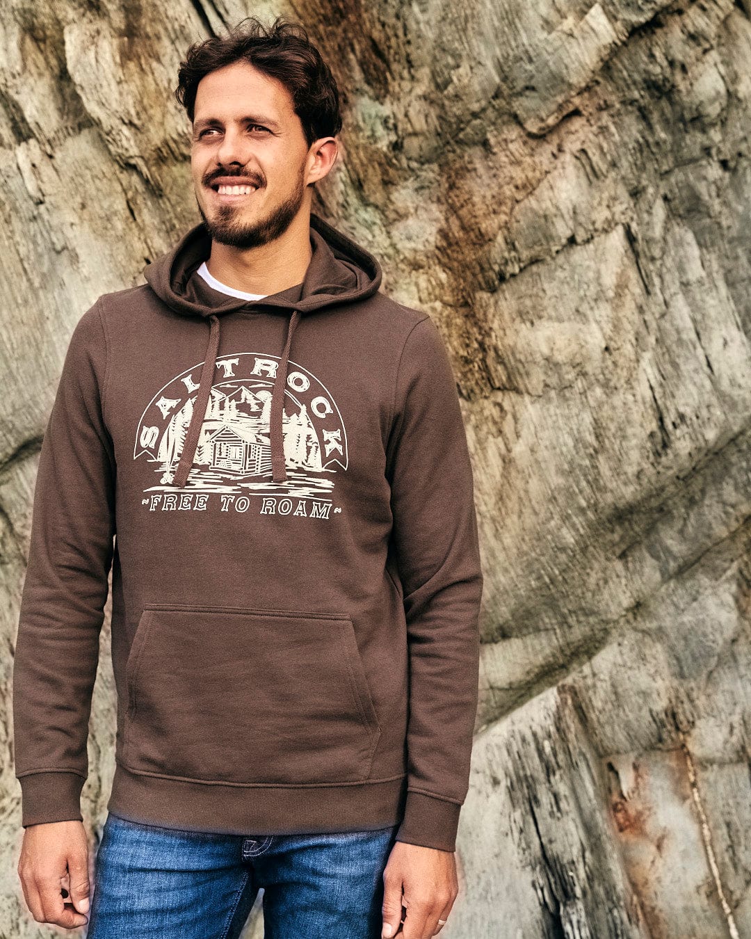 A man wearing a Saltrock Camping Cabin - Mens Pop Hoodie - Brown, with drawstring hood, standing in front of a rock.