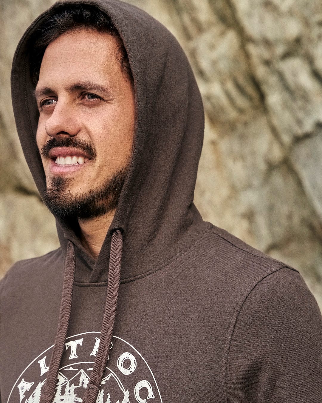 A man in a Saltrock Camping Cabin - Mens Pop Hoodie - Brown with a drawstring hood smiling in front of a rock.