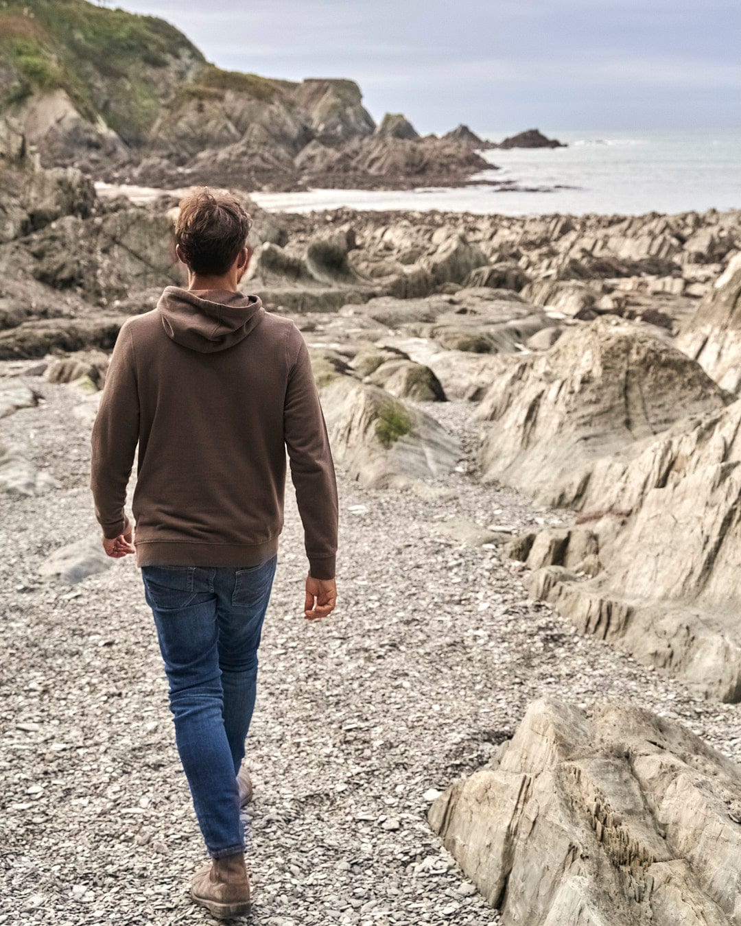 A man wearing a Saltrock Camping Cabin - Mens Pop Hoodie - Brown is walking down a rocky path towards the ocean.