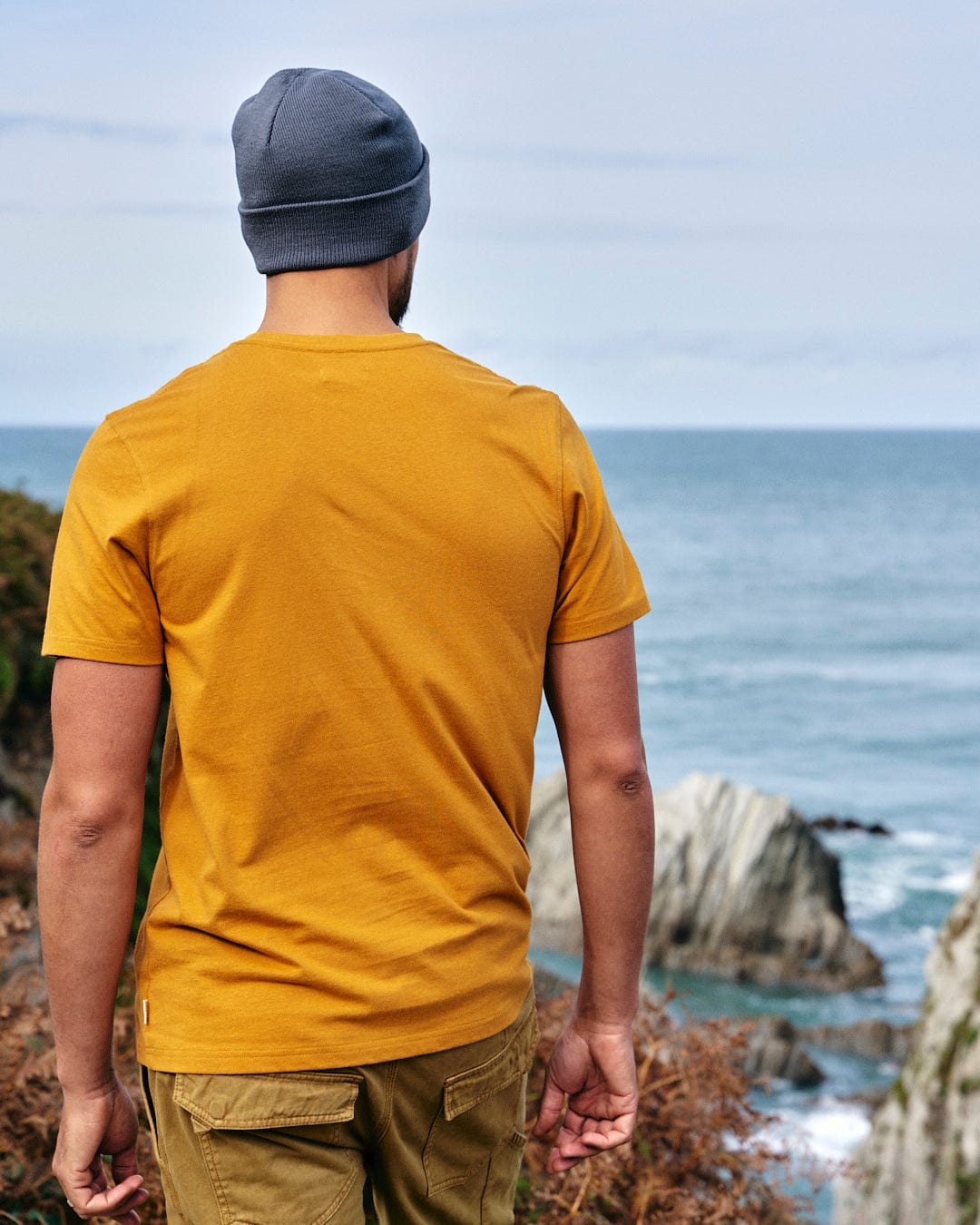 A man wearing a yellow Saltrock Camping Cabin T-Shirt looking at the ocean.