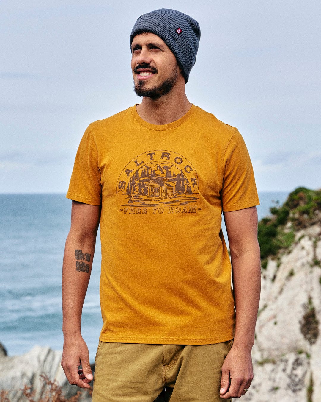 A man wearing a Saltrock Camping Cabin - Mens Short Sleeve T-Shirt - Yellow in front of the ocean.