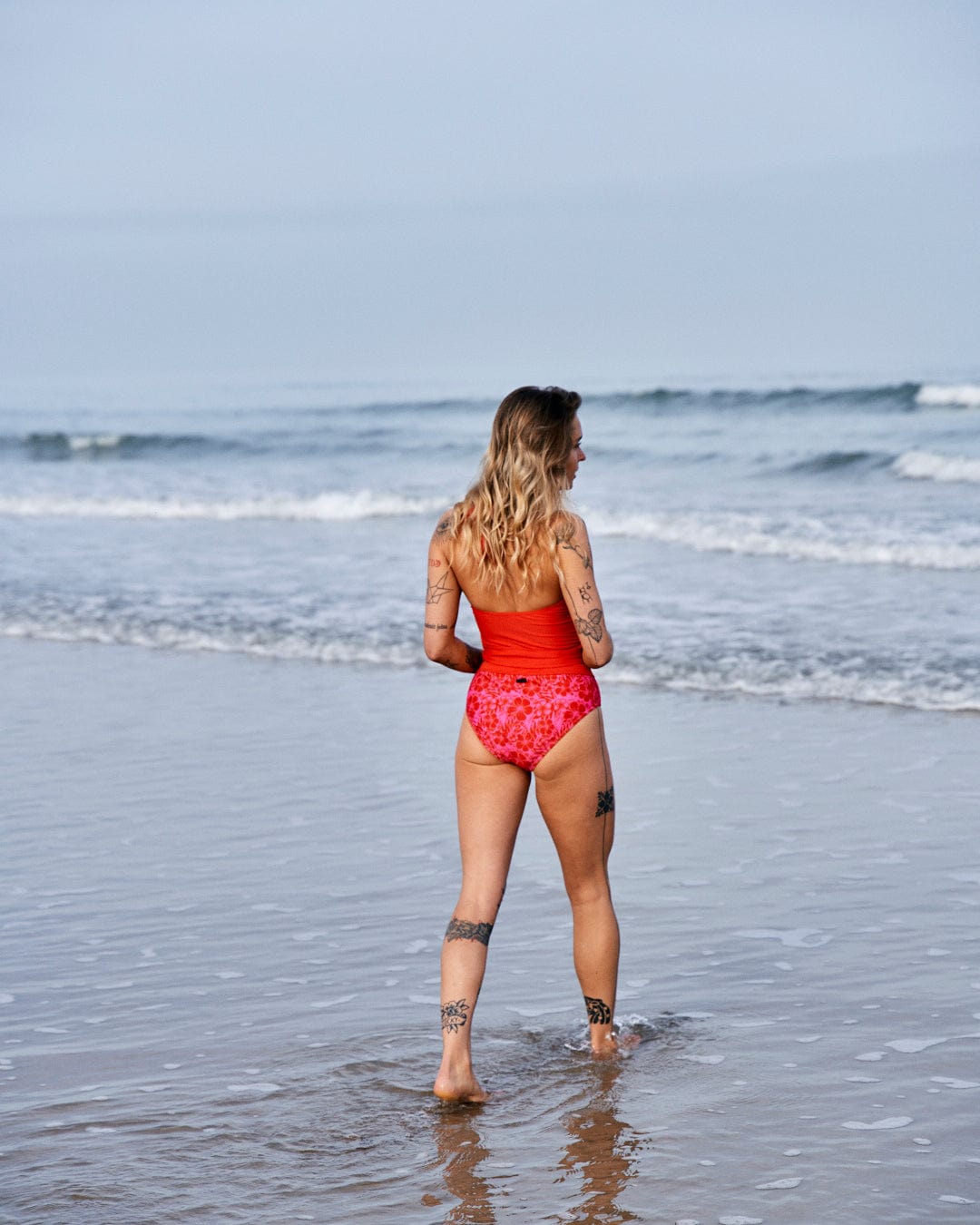 Woman in Saltrock's Carly Hibiscus - Recycled Womens Halter Neck Bikini Top - Pink standing in the ocean, looking out at the waves, with visible tattoos on her arms and legs.