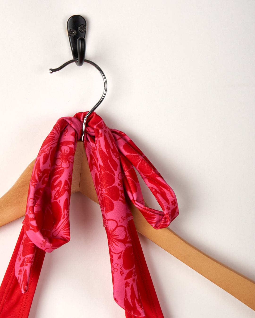 A Carly Hibiscus - Recycled Womens Halter Neck Bikini Top in Pink, made from recycled fibers, hangs from a wooden hanger, attached to a black wall hook on a white background. (Saltrock)