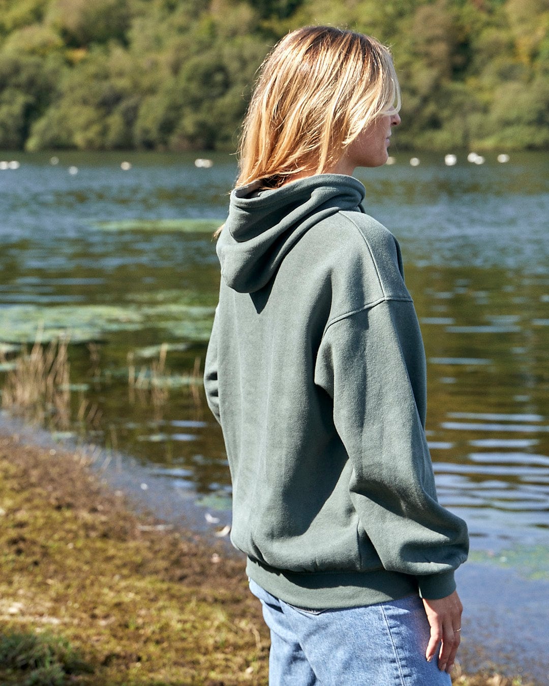 A woman wearing a Saltrock Celeste - Womens Pop Hoodie - Dark Green stands next to a body of water.