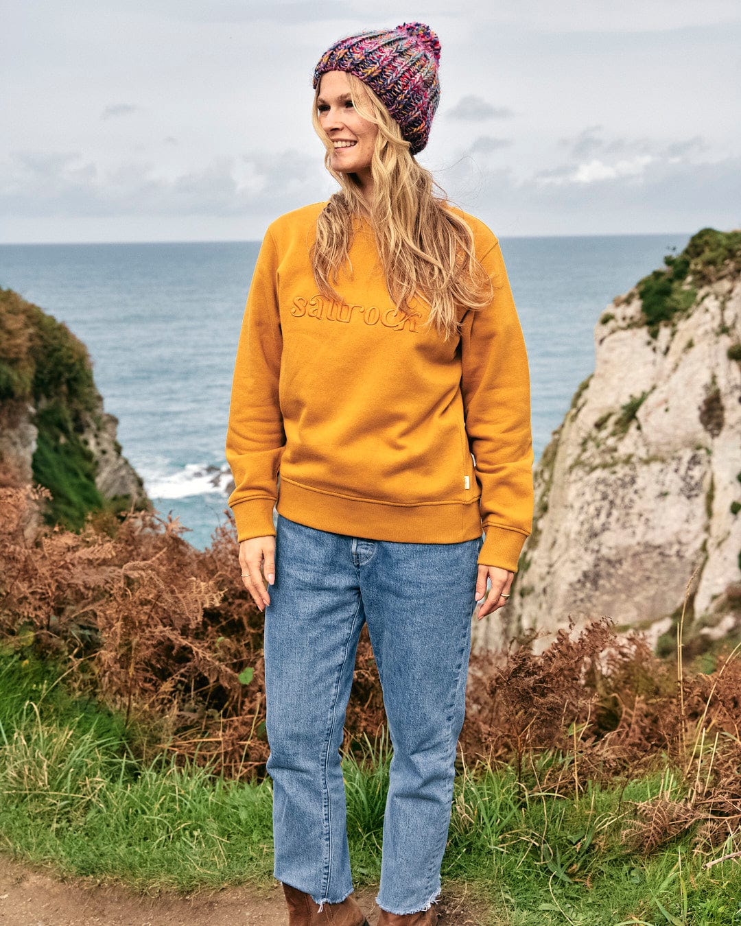 A woman wearing a yellow Saltrock Celeste - Womens Sweat - Yellow sweatshirt standing on a path near the ocean.