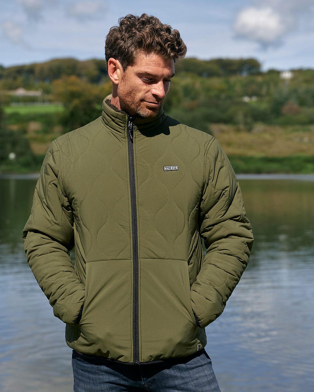 A man is standing by a lake wearing a Saltrock Cirrus - Quilted Reversible Jacket - Dark Green.