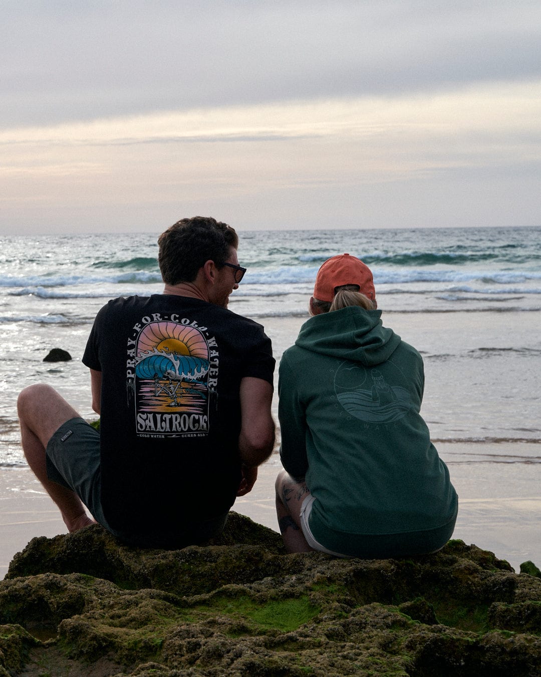 Two people wearing the Coldwater Club - Ladies Zip Hoodie in Green by Saltrock sit on a rocky beach, looking out at the ocean.