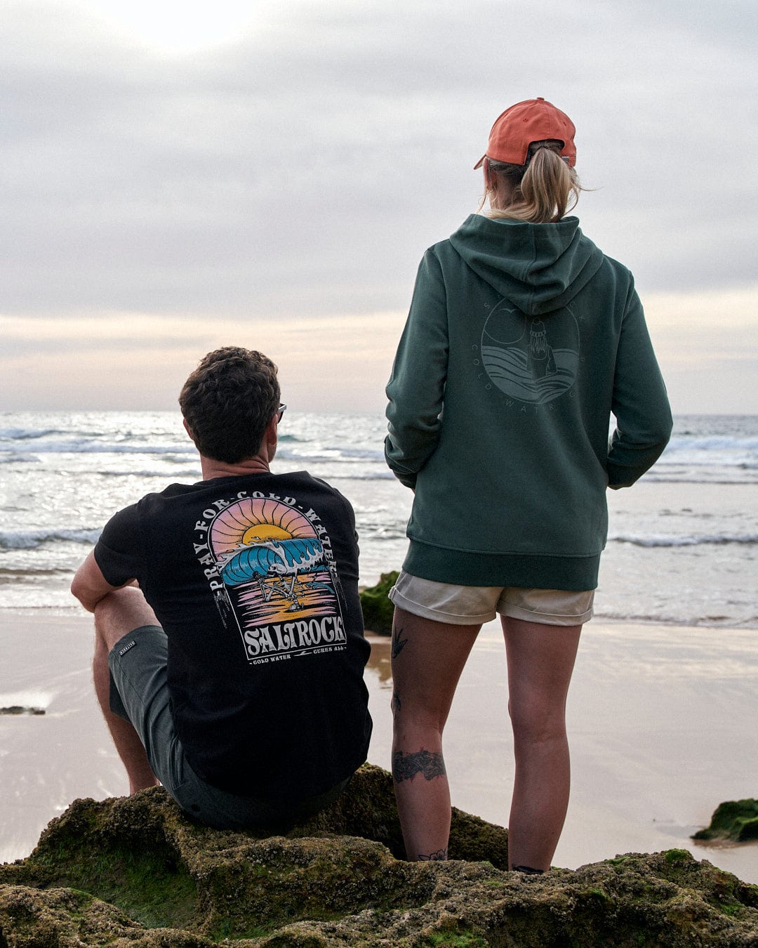 Two individuals clad in Saltrock's Coldwater Club - Ladies Zip Hoodie - Green standing on a rocky shore, gazing out at the sea.