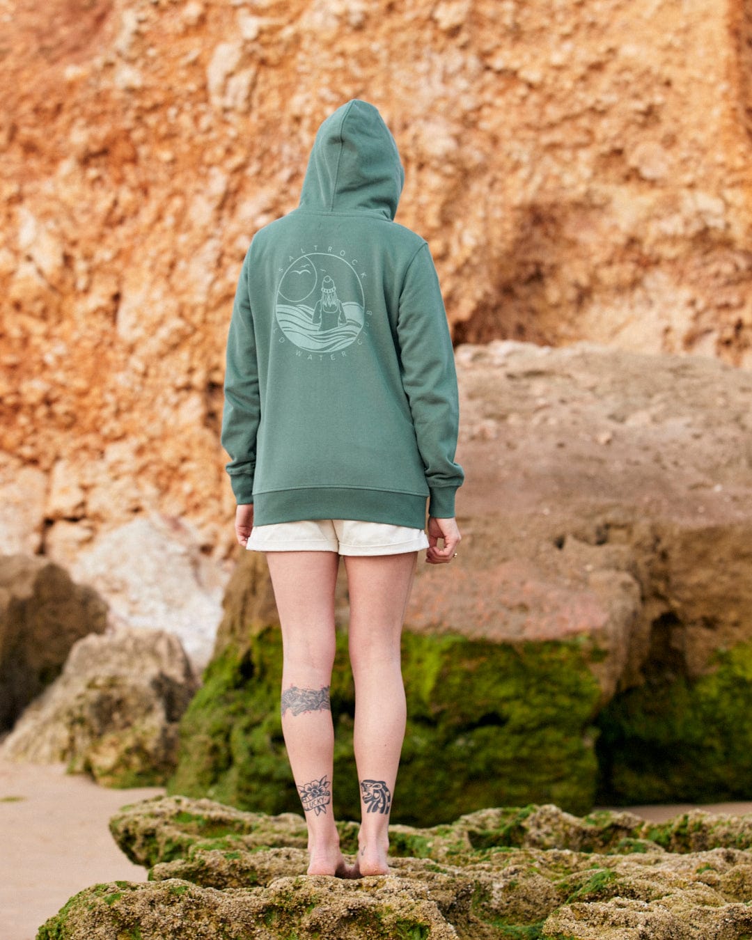 A person in a Coldwater Club - Ladies Zip Hoodie - Green standing on a rocky beach facing away from the camera, featuring subtle Saltrock branding.