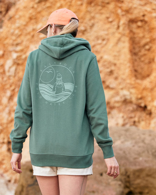 Woman in Coldwater Club - Ladies Zip Hoodie - Green with Saltrock branding and beige shorts standing in front of a rocky background.
