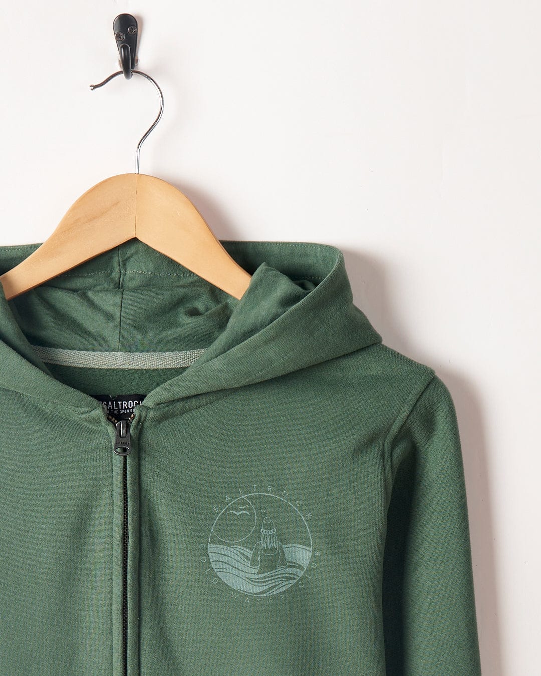 A Coldwater Club - Ladies Zip Hoodie - Green with a front zipper hanging on a wooden hanger against a white background, featuring a circular Saltrock branding logo on the left chest area.