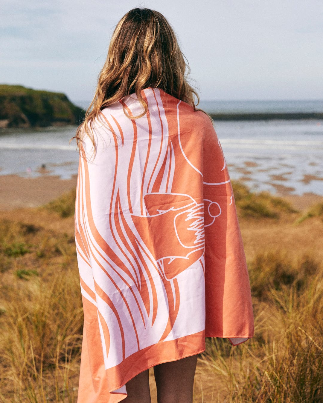 Woman standing on a beach, facing the sea, with a Cold Water Club - Microfibre Towel - Orange towel draped over her shoulders.