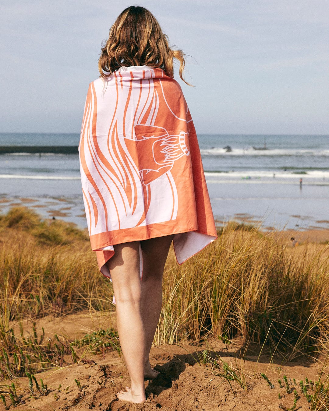 Woman draped in a Saltrock Cold Water Club Microfibre Towel in Orange and White stripes stands on a sandy beach, overlooking the ocean with surfers in the distance.