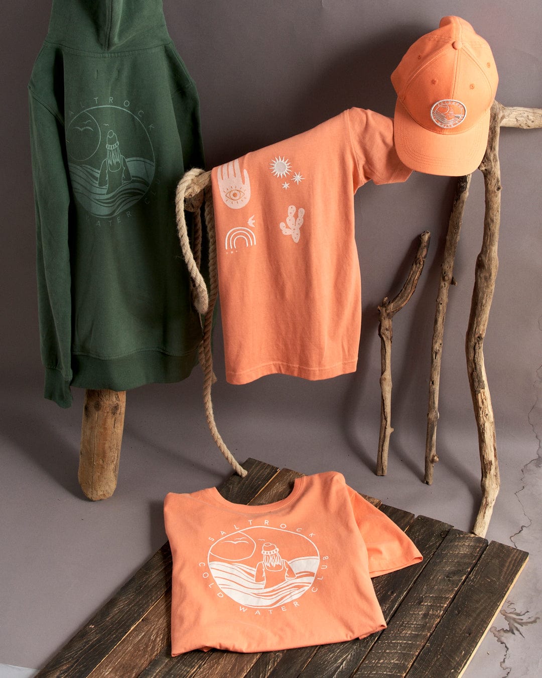 A collection of adventure-themed clothing and accessories displayed on wooden branches, featuring a Saltrock Coldwater Club green cotton hoodie, an orange cotton t-shirt, and a matching cap.