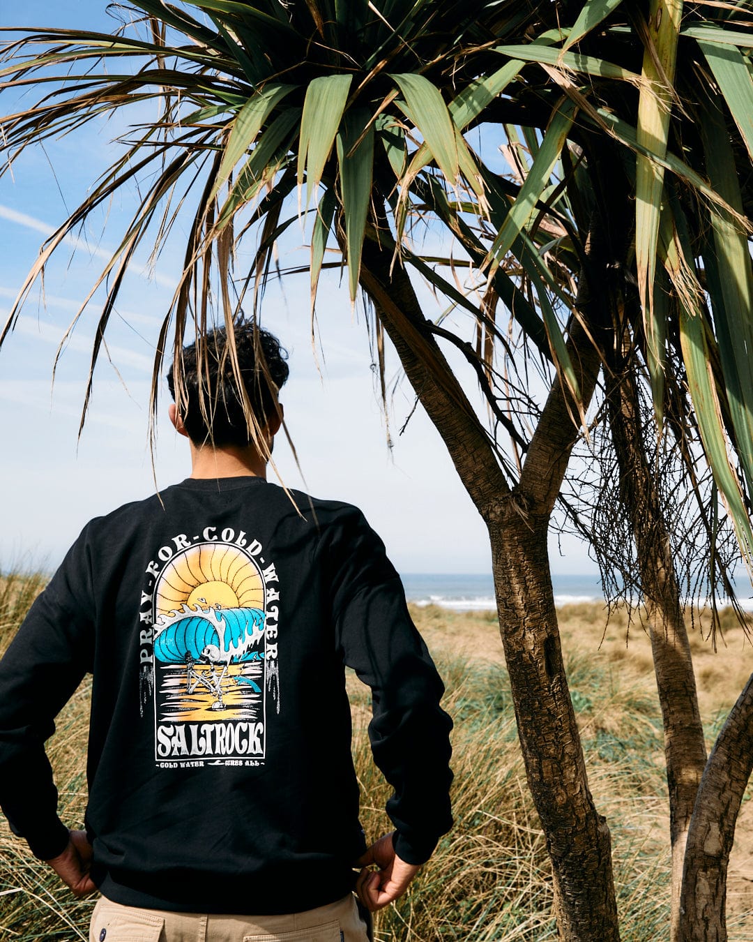 A person wearing a Saltrock Cold Water - Mens Oversized Sweat - Black stands facing the ocean, partially obscured by a palm tree in the foreground.