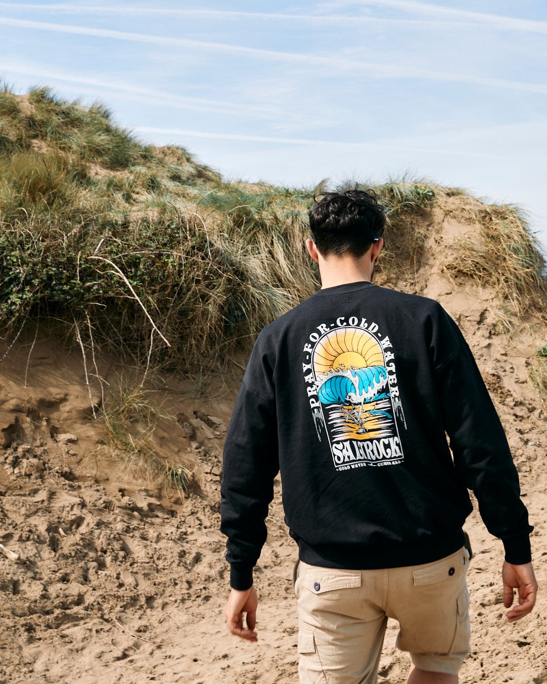 A man in a black Saltrock Cold Water - Mens Sweat hoodie and khaki shorts viewing sand dunes, with his back to the camera.