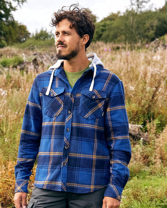 A man in a Saltrock Colter - Mens Hooded Shirt in Blue plaid, made from high quality fabric, standing in a field.