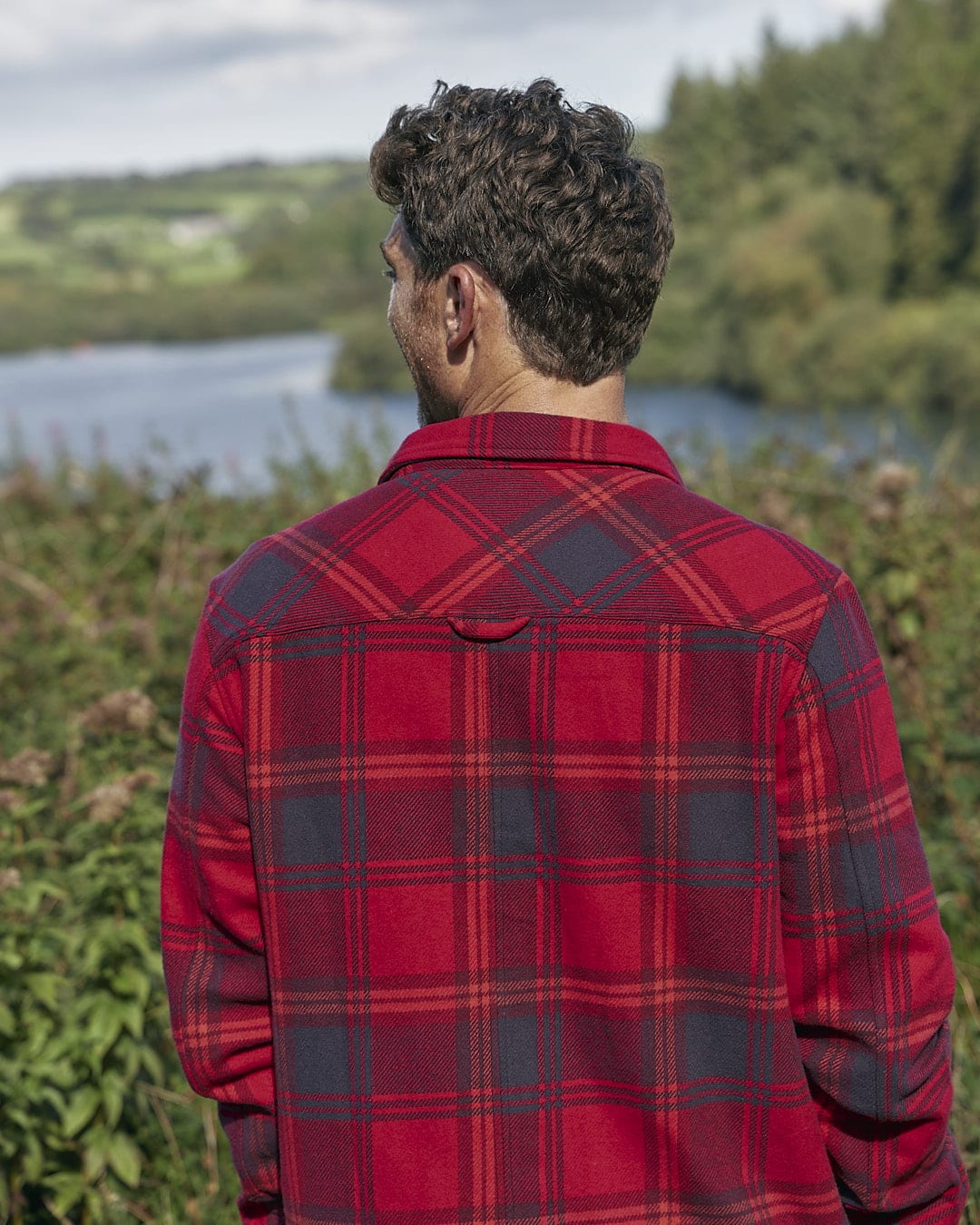 A man wearing a red Saltrock Colter Hooded Shirt looking at a lake.