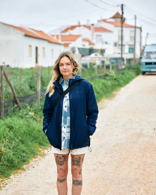 A woman with tattoos on her legs stands on a rural road, wearing a Saltrock Cooper - Womens Packable Waterproof Jacket in Blue and shorts, with houses and a bus in the background.
