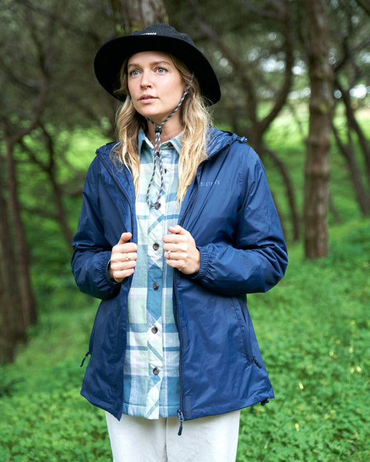 A woman in a Saltrock Cooper - Womens Packable Waterproof Jacket in blue and black hat stands in a lush green forest, looking slightly to the left with a thoughtful expression.