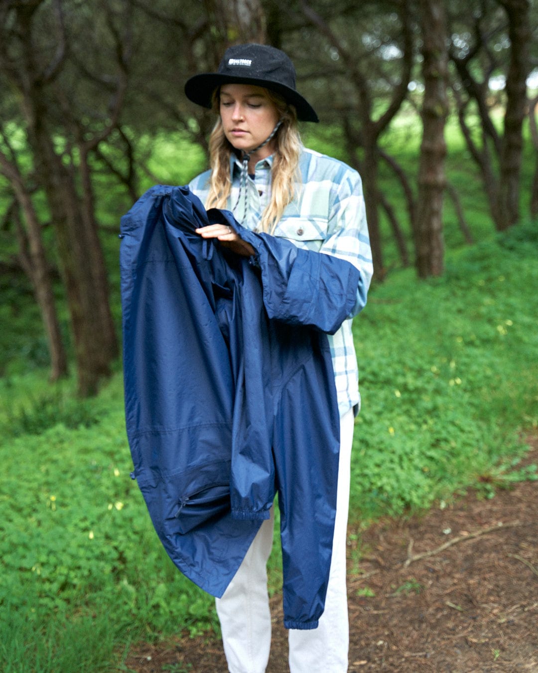 Woman in a bucket hat and plaid jacket folding a Saltrock Cooper - Womens Packable Waterproof Jacket in Blue in a lush green forest.