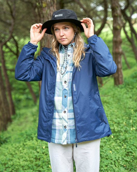 Woman in a Saltrock Cooper - Womens Packable Waterproof Jacket - Blue and black hat standing in a lush green forest, adjusting her hat and looking at the camera.