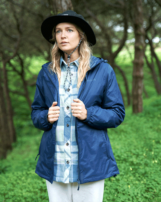 Woman in a Saltrock Cooper - Womens Packable Waterproof Jacket in Blue and black hat standing in a green forest, looking slightly to the side.