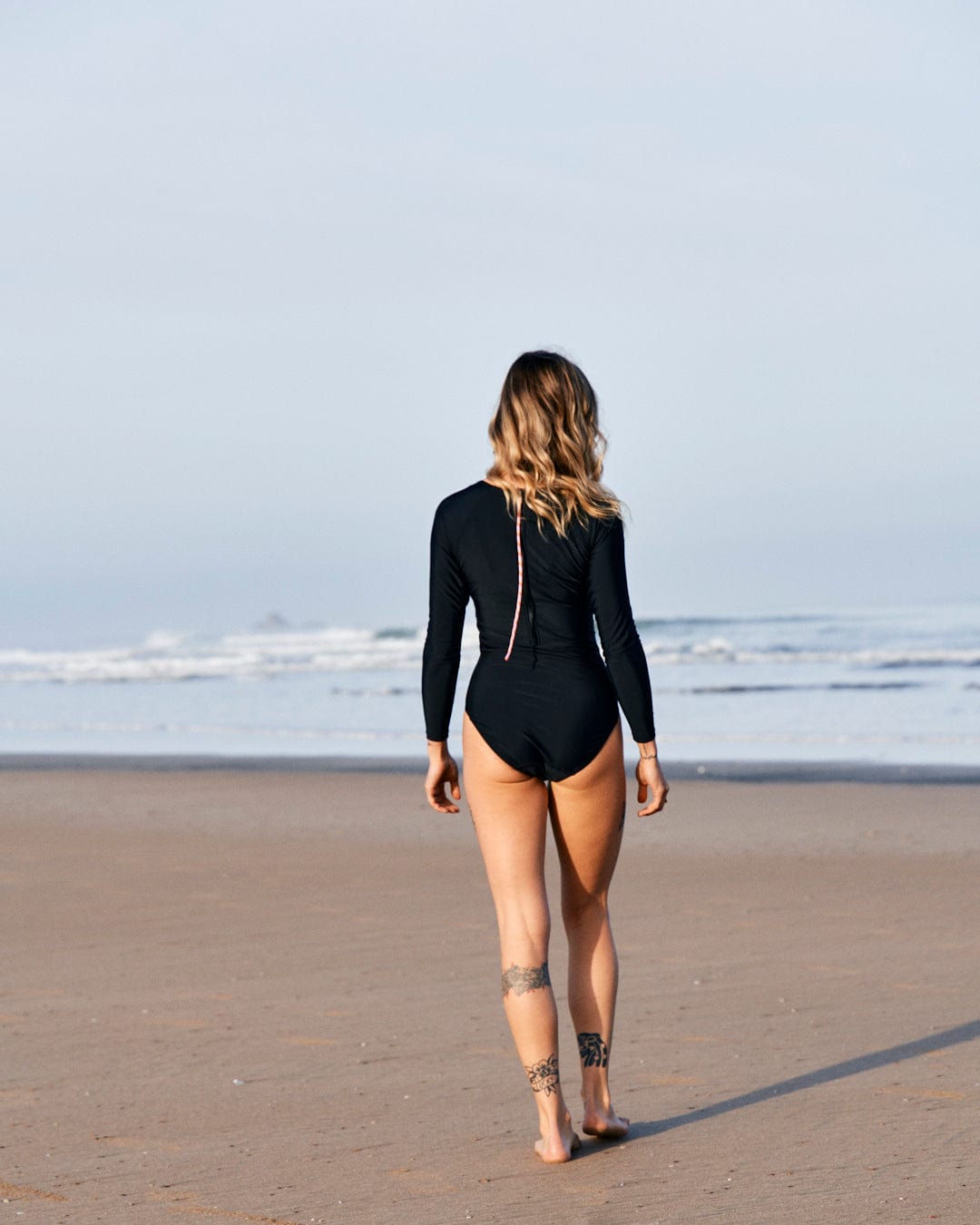 A woman in a Saltrock Cora Retro - Recycled Womens Long Sleeve Swimsuit - Dark Grey walks toward the ocean on a beach, viewed from behind, with a tattoo visible on her left thigh.