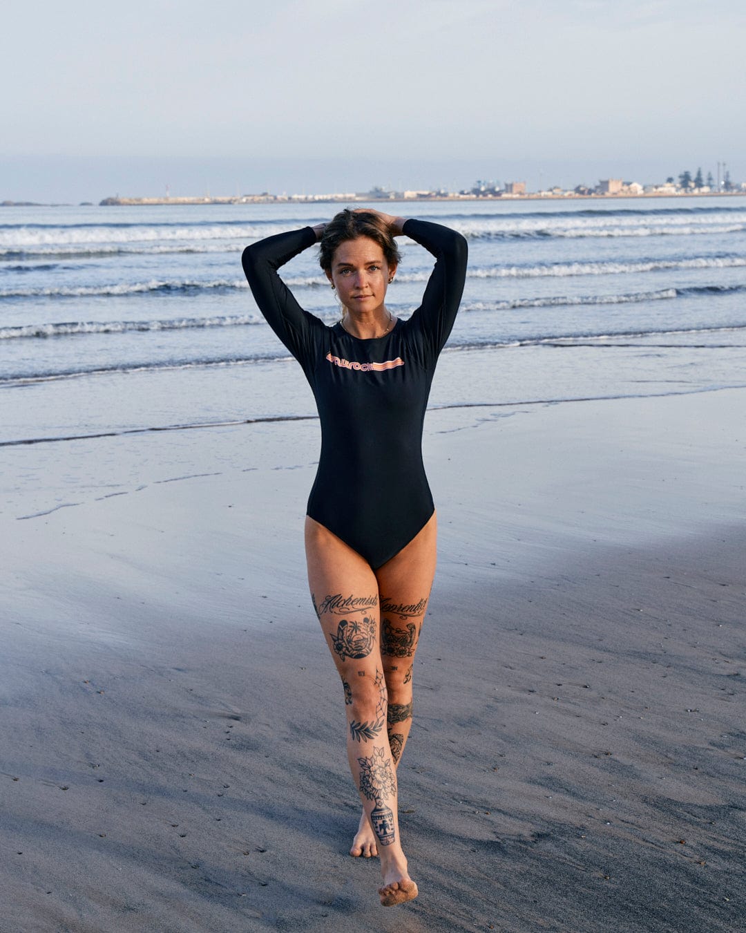 A woman in a Saltrock Cora Retro recycled women's long sleeve swimsuit in dark grey stands on a beach, hands on her head, with visible tattoos on her legs and an industrial backdrop.