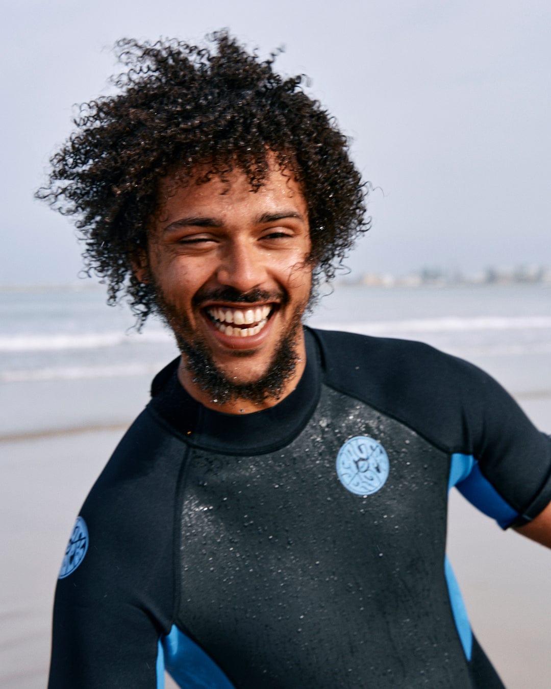 A person with curly hair and a beard smiles while wearing a Saltrock Core - Mens 3/2 Shortie Wetsuit - Black/Blue on a beach. The ocean and sky are visible in the background.