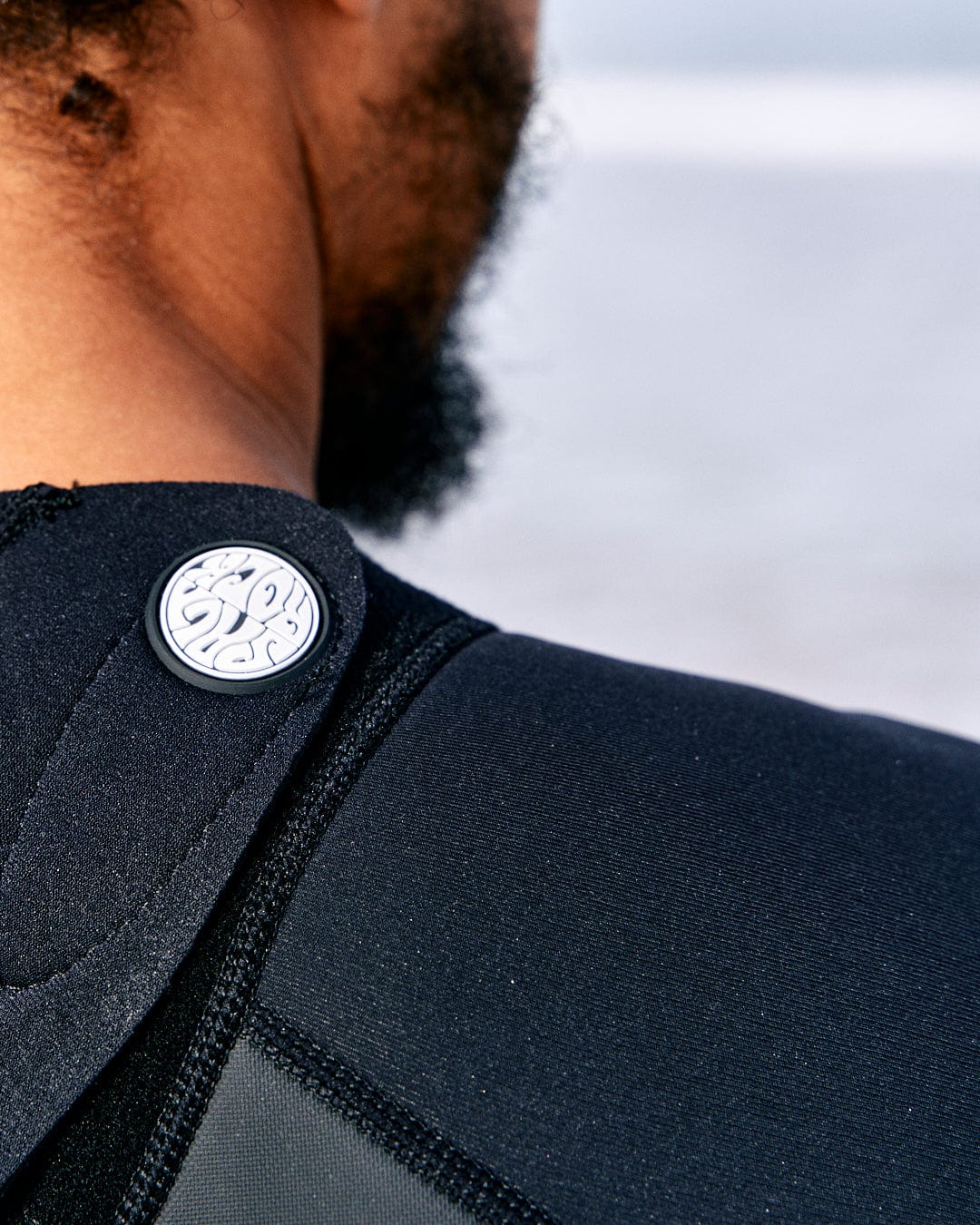 Close-up of a person's neck and shoulder wearing a Saltrock Core - Mens 3/2 Shortie Wetsuit - Black/Blue, featuring a circular emblem with a stylized wave design on the back collar.