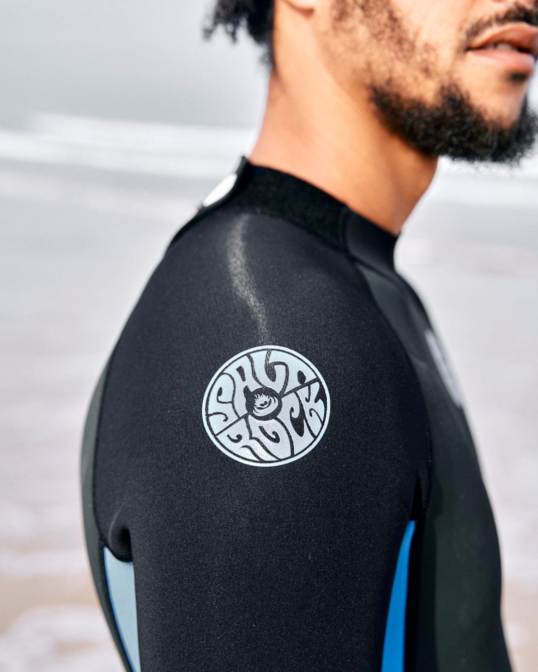 Close-up of a person wearing a black neoprene/nylon wetsuit standing on a beach. The sleeve has a circular logo with stylized text. The 3/2mm double line material is visible, and the background shows a blurry view of the ocean and sand. They are wearing the Core - Mens 3/2 Shortie Wetsuit - Black/Blue from Saltrock.