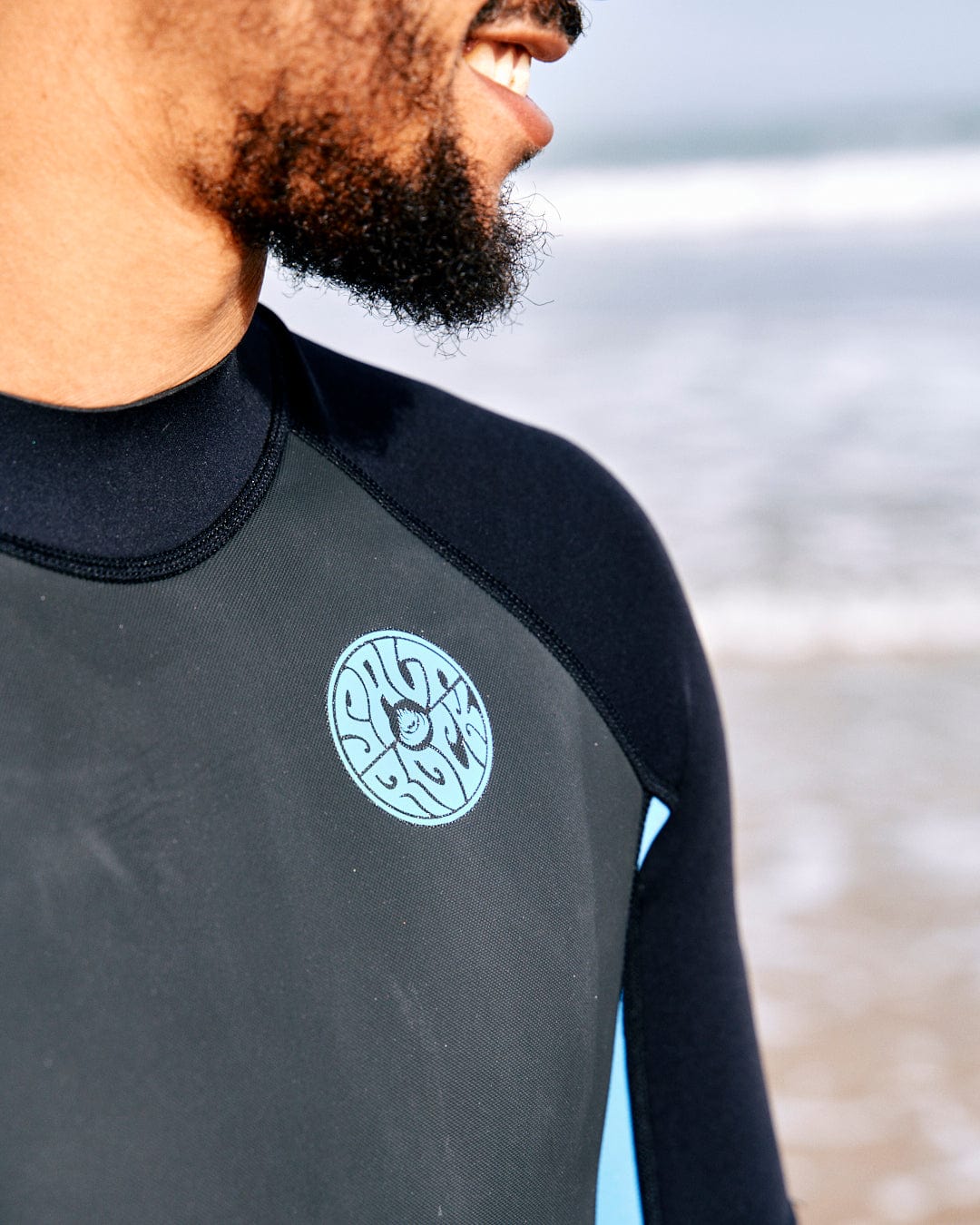 A person wearing a black neoprene Core - Mens 3/2 Shortie Wetsuit - Black/Blue by Saltrock with a circular logo on the mesh skin chest stands by a sandy beach with waves in the background.