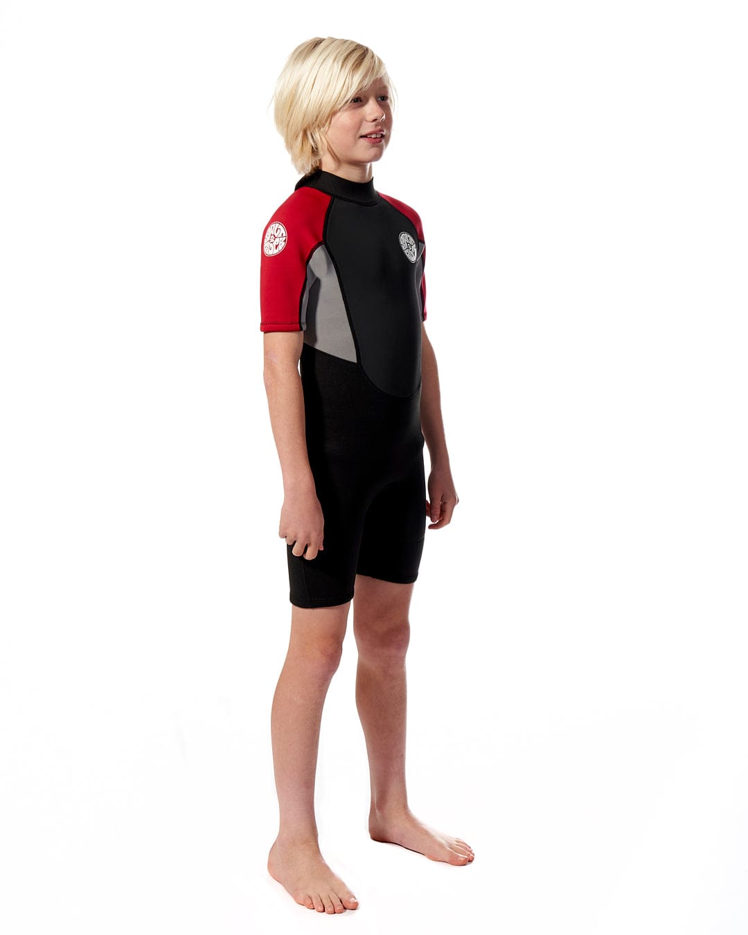 A young boy in a Saltrock Core - 3/2 Shortie Wetsuit - Red standing in front of a white background.