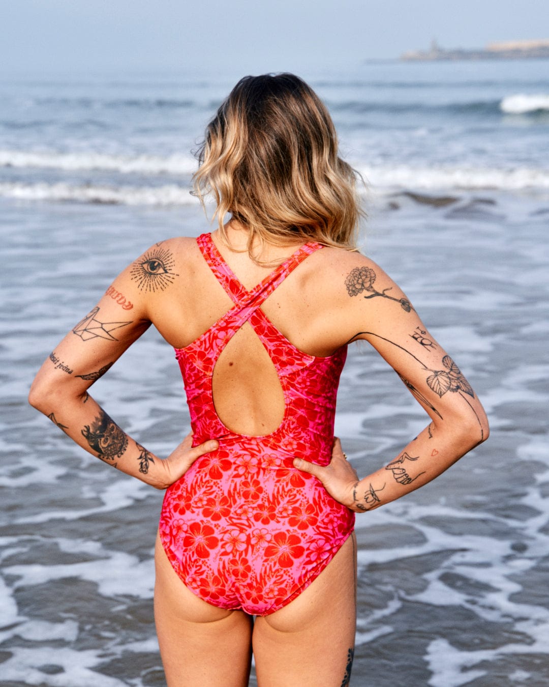 A woman in a red full piece swimsuit with Corrine Hibiscus print and visible tattoos stands on a beach, looking towards the sea.