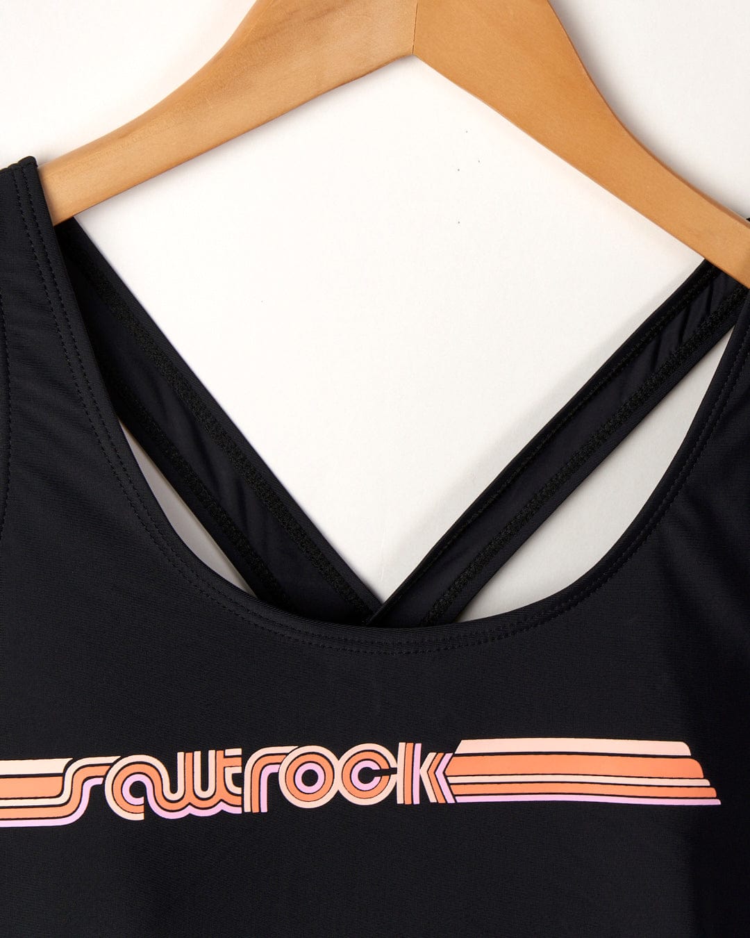 Close-up of a Corrine Retro swimsuit in black made from recycled fibers with the word "Saltrock" and three horizontal stripes in pink and orange, displayed on a hanger against a white background.