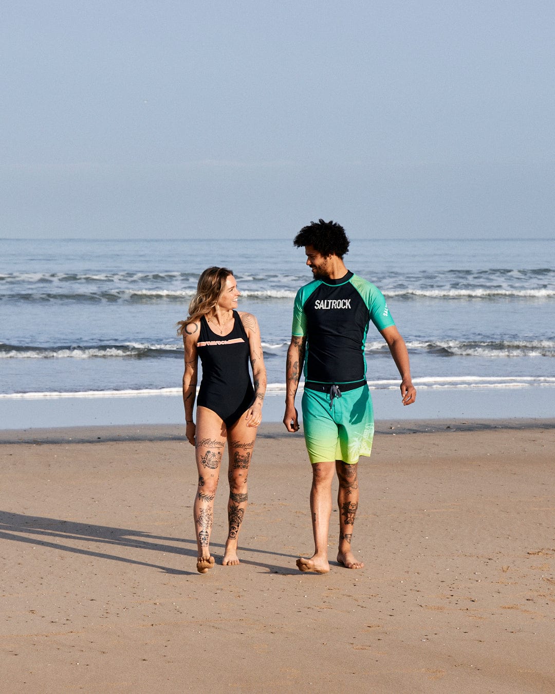 A man and a woman walking on a beach, discussing, both dressed in swimwear including a Corrine Retro - Recycled Womens Swimsuit in Black by Saltrock, with clear skies and calm sea in the background.