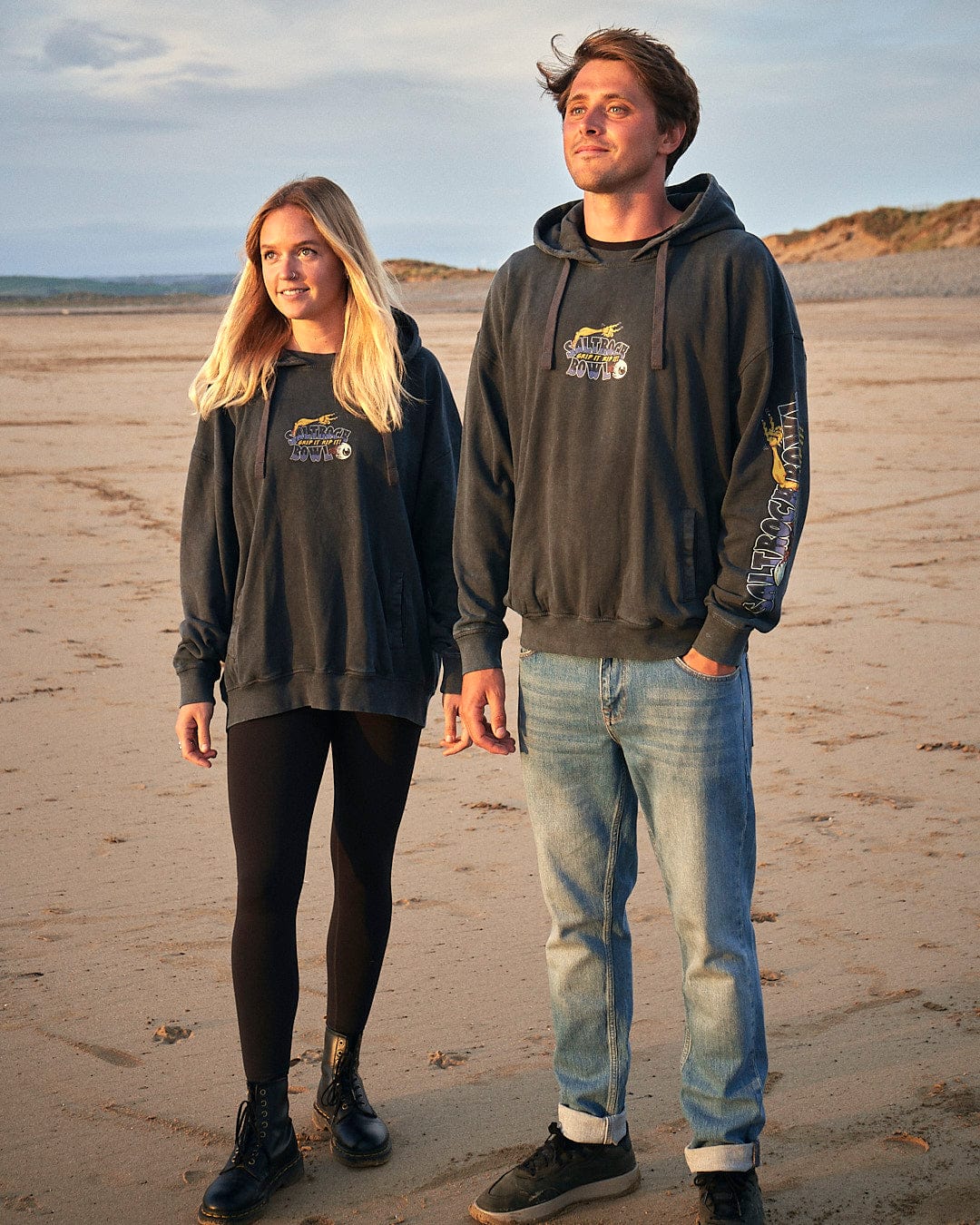 A man and woman, both donning stylish Creeper Encore - Mens Pop Hoodie - Washed Black hoodies, stand side by side on a serene beach. The hoodies feature Saltrock branding, showcasing their quality and trendy design. The couple.