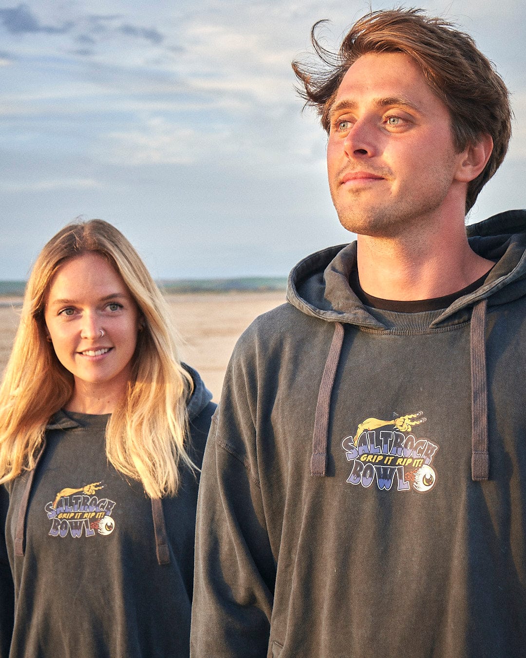 A man and woman wearing Saltrock Creeper Encore - Mens Pop Hoodie - Washed Black stand relaxed on a beach.