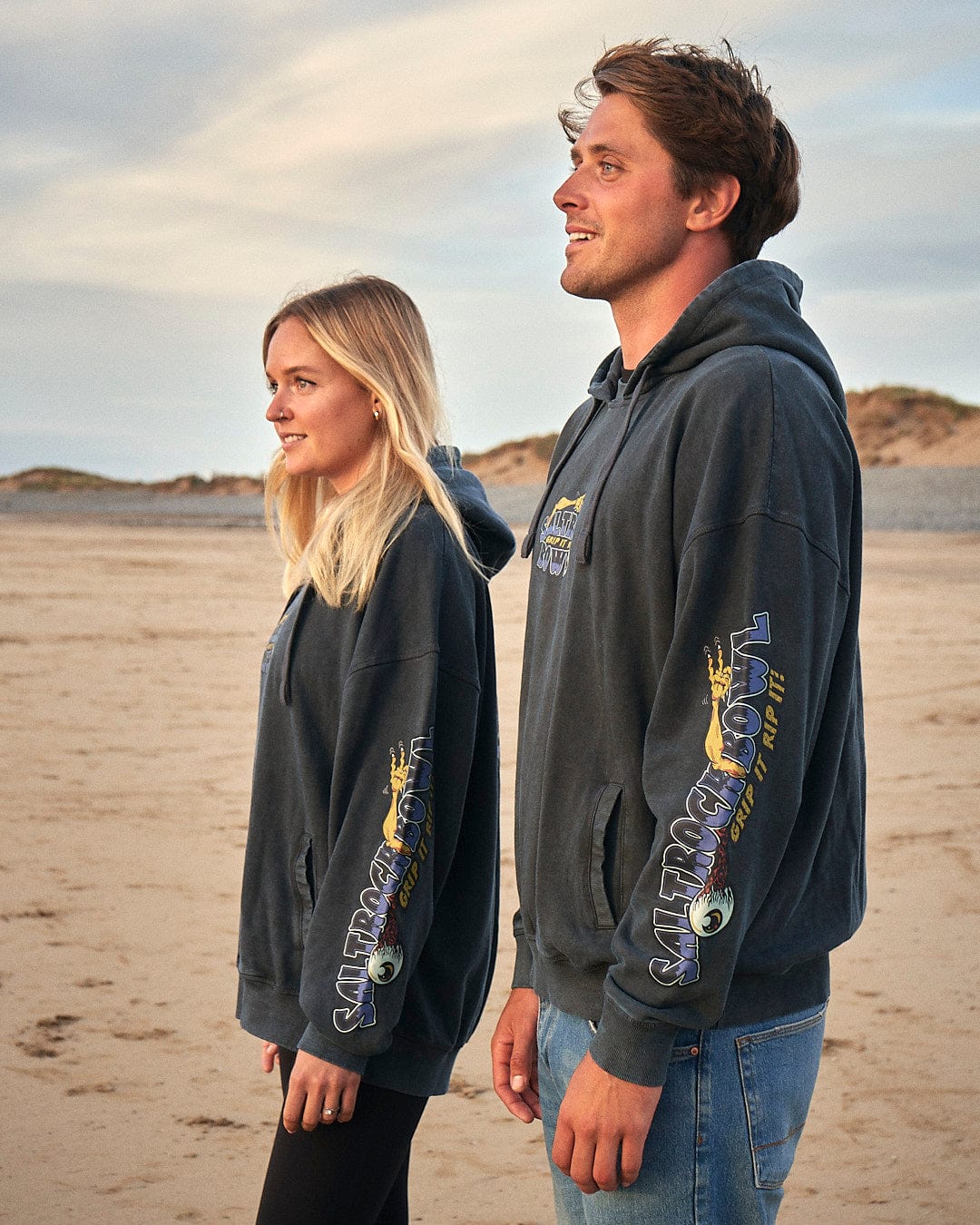 A man and woman standing on a beach wearing Saltrock brand Creeper Encore - Mens Pop Hoodie - Washed Black hoodies.