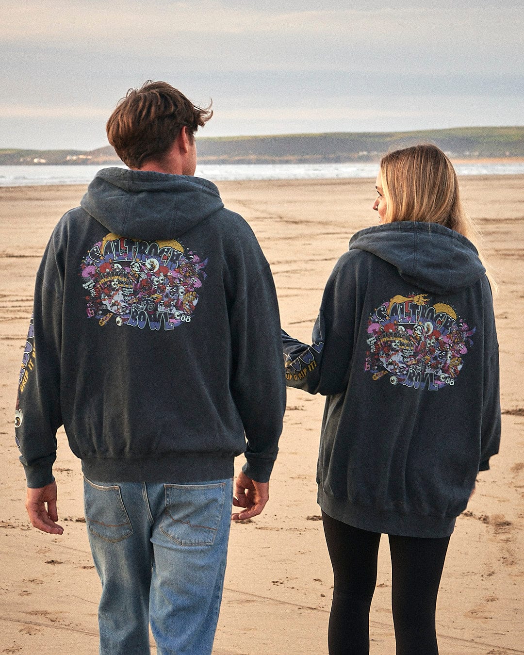 Two people walking on a beach wearing Saltrock Creeper Encore - Mens Pop Hoodie - Washed Black.