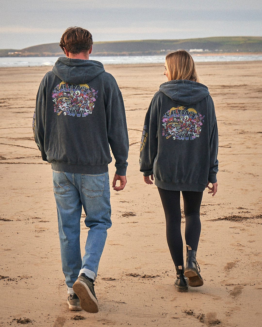 Two people wearing Saltrock Creeper Encore - Mens Pop Hoodie - Washed Black hoodies stroll along the beach, showcasing the trendy graphic print designs.